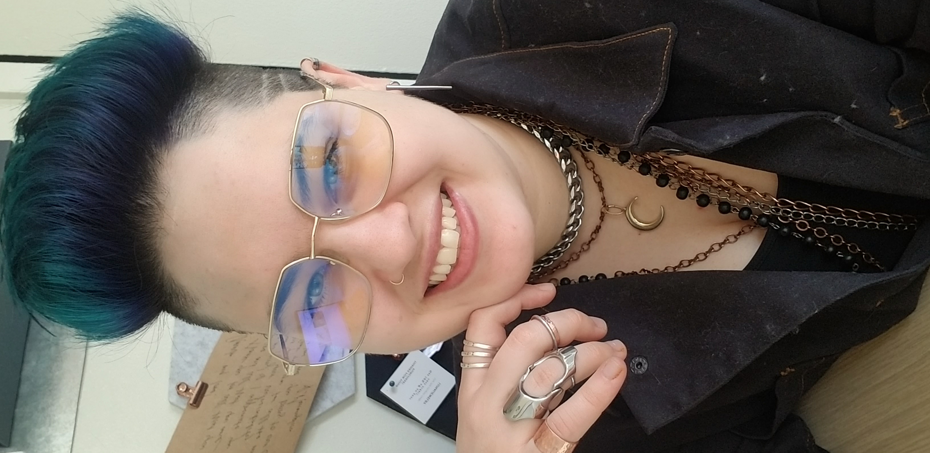 Photo of Ara Phoenixx, a nonbinary human with blue hair, glasses, and a variety of jewelry. Ara wears a dark jumpsuit, is sitting at a desk, and is smiling.